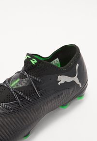 Scarpa da calcio nera con superficie texturizzata, dettagli verdi e logo Puma riflettente. Presenta lacci neri e un design aderente e a profilo basso.