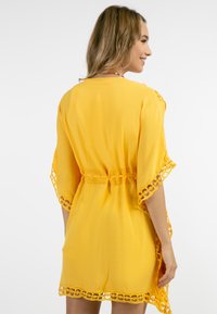 Keltainen kimono-tyylinen peite, jossa on pitsireunus, lyhyet hihat ja vyötäröltä tiukennettu malli. Kevyt kangas, jossa on laskostuva siluetti.