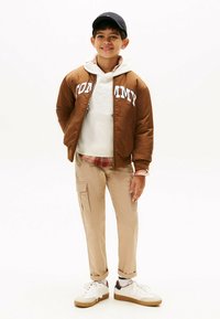 Bruine bomberjack met witte "TOMMY" logo, witte hoodie en tan cargobroek. Witte sneakers met bruine accenten. Casual gelaagd outfit.