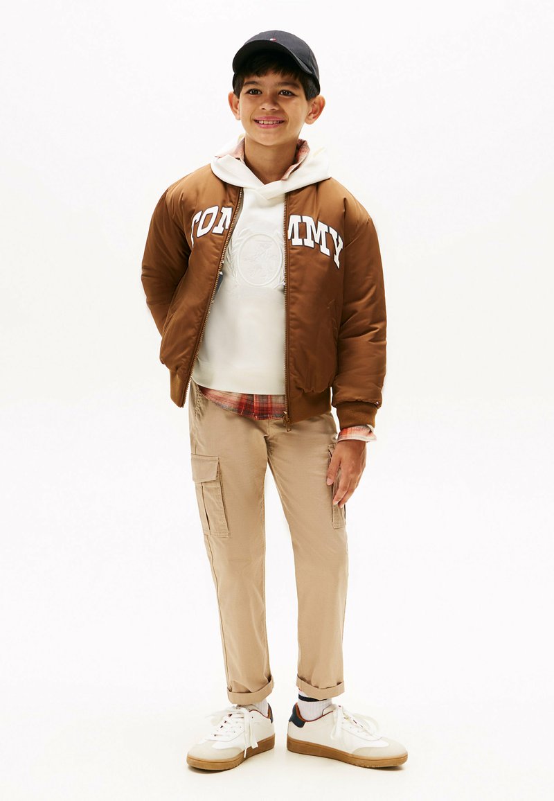 Bruine bomberjack met witte "TOMMY" logo, witte hoodie en tan cargobroek. Witte sneakers met bruine accenten. Casual gelaagd outfit.
