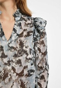 Blouse transparente présentant un motif semblable à de l'aquarelle dans des tons de bleu, de gris et de marron, avec un col volanté et de subtiles rayures verticales.