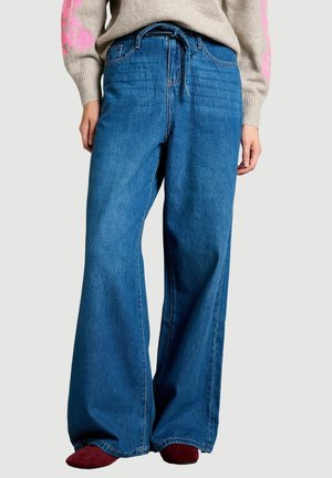 Mujer con vaqueros azules de pierna ancha y cintura con cordón, y zapatos rojos, combinados con un suéter beige con mangas florales rosas.