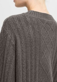 Grauer Strickpullover mit strukturiertem Muster, das diagonale Rippungen und Rautenoptimierungen enthält. Ribbelkragen und weiches, dickes Material.
