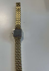 Reloj cuadrado de oro con una correa de metal texturizada. La esfera es metálica con una superficie reflectante y un logotipo grabado en la parte posterior.