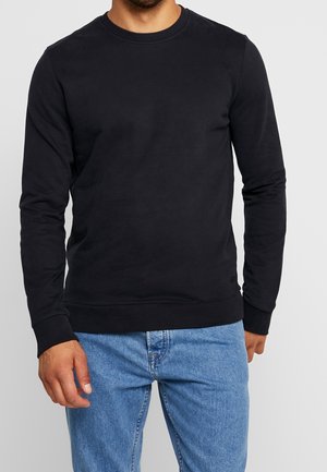 Mand iført en ensfarvet sort langærmet sweatshirt med rund hals og lyseblå jeans, stående foran en grå baggrund.