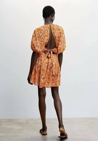Robe fleurie orange avec des manches courtes bouffantes, dos nu et un détail à nouer à la taille. Tissu léger avec une silhouette fluide.