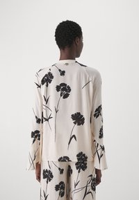 Persona che indossa un top a maniche lunghe color crema e pantaloni con stampe floreali nere, vista da dietro su uno sfondo semplice.