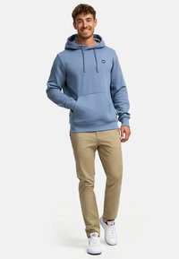 Lys blå hettegenser med justerbare snorer og frontlomme, parret med beige bukser og hvite sneakers. Ryddig, avslappet stil.