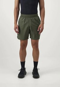 Hombre vestido con pantalones cortos de cintura elástica verdes oliva, camiseta sin mangas negra, calcetas cortas negras y zapatillas deportivas negras, de pie sobre un fondo liso.