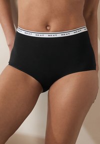 Sous-vêtement noir en coton taille haute avec une ceinture élastique blanche arborant le nom de la marque "NEXT" en texte noir. Texture lisse.