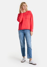 Blonde Frau, die ein rotes Langarmoberteil mit einer Brusttasche, blaue Jeans und weiße Sneaker trägt, steht und schaut zur Seite.