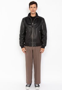 Schwarze Leder-Bomberjacke mit Stehkragen, Reißverschluss vorne und Seitentaschen, kombiniert mit braunen Hosen und schwarzen Sneakers.