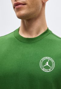 Κοντινό πλάνο ενός ατόμου που φοράει πράσινο μπλουζάκι με λευκό λογότυπο Jordan Golf στο στήθος, δείχνοντας μέρος του λαιμού και των χειλιών.