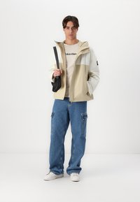 Jachetă color-block în bej deschis și alb, tricou casual kaki, pantaloni cargo din denim albastru, purtând o geantă de umăr neagră, încălțat cu pantofi sport albi.