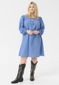 Vestido azul texturizado con escote con cordones y mangas abullonadas, combinado con botas vaqueras negras con acentos plateados.