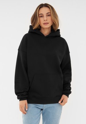 Felpa nera con cappuccio, spalle scese e tasca frontale. Tessuto morbido, vestibilità rilassata, polsini e bordo a costine. Indossata con jeans azzurri chiari.