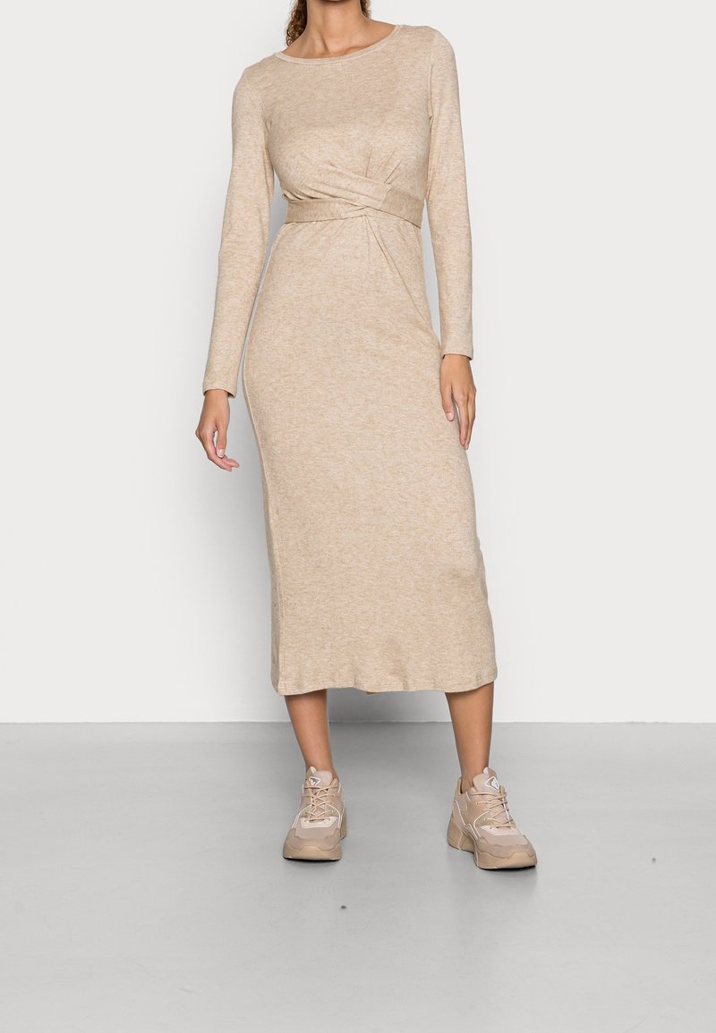 Frau in einem beigen, langärmligen Midi-Strickkleid mit einer Taillenfaltendetail, kombiniert mit hellbeigen klobigen Turnschuhen, steht auf einem grauen Boden.