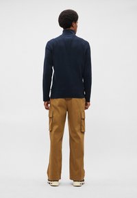 Σκούρο μπλε πλεκτό πουλόβερ με ψηλό γιακά, συνδυασμένο με καφέ cargo παντελόνι που διαθέτει πλάγιες τσέπες και άνετη εφαρμογή, φορεμένο με ελαφριά αθλητικά παπούτσια.