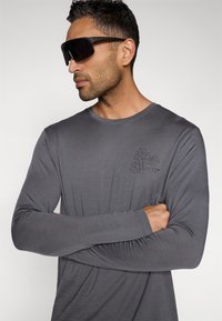 Grå langermet skjorte laget av glatt stoff med et blomsterdesign i svart på venstre brystområde. Modellen har på seg mørke solbriller.