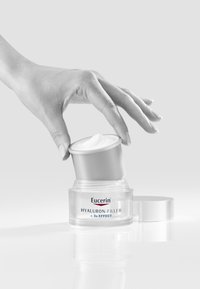 Eucerin Hyaluron-Filler cream in a clear jar with a silver cap. A hand holds the cap, revealing a smooth white cream inside.