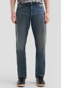 Jeans in denim blu con vestibilità dritta, caratterizzati da un leggero sbiadimento, cinque tasche e un classico cinturino con passanti. Indossati con scarpe da ginnastica nere.