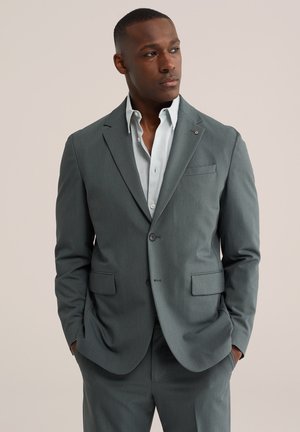 Mies, jolla on harmaa pukutakki ja housut, valkoinen paita, kädet taskuissa, katsoo oikealle yksivärisellä beige taustalla.