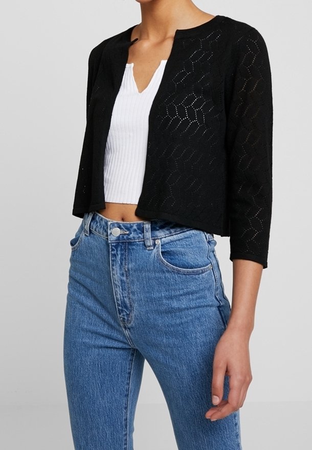 Svart cardigan med trekvartslånga ärmar och ett texturerat, öppet stickat mönster, bärs över en vit tajt topp och kombineras med högmidjade blå jeans.