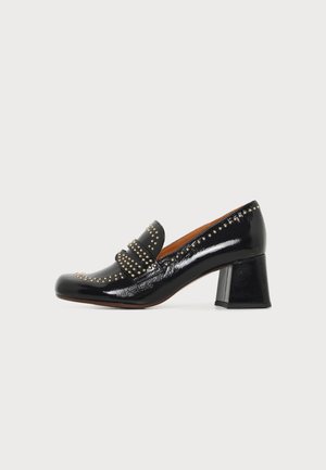 Schwarzer Lacklederschuh mit niedrigem Absatz, goldenen Nieten entlang der Kanten und des Riemens, Blockabsatz und abgerundeter Spitze auf weißem Hintergrund.
