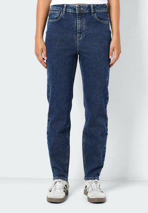 Personne portant un jean droit taille mi-haute bleu foncé et des baskets blanches, mains détendues le long du corps sur un fond uni.