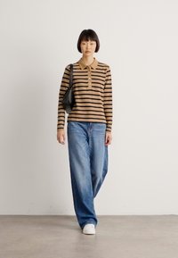 Donna che cammina, indossando una polo a righe lunga color beige e nera, jeans blu a gamba larga, sneakers bianche, borsa a tracolla nera, su uno sfondo bianco semplice.