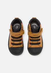 Zapatillas de cuero marrón y negro con una correa de velcro, cordones negros, suelas de goma y acentos rojos. Presentan una puntera texturizada y un cuello acolchado.
