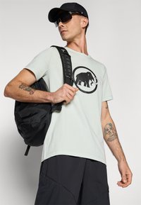 Light green t-shirt with a black elephant graphic, black backpack, black cap, and dark pants; features visible tattoos on arm.