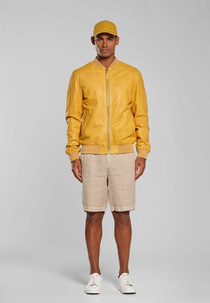 Mężczyzna stojący, ubrany w żółtą czapkę z daszkiem, żółtą kurtkę zapinaną na zamek, beżowe szorty i białe trampki, na jednolitym jasnoszarym tle.