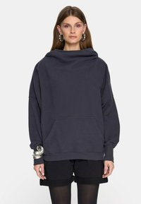 Kobieta w ciemnym, oversizedowym hoodie z przednią kieszenią, czarnych szortach, czarnych rajstopach, dużej srebrnej bransoletce oraz kolczykach w kształcie koła na jednolitym tle.