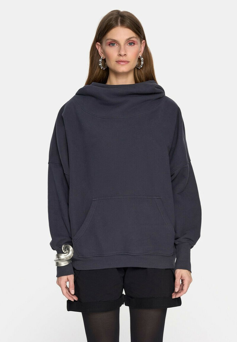 Kobieta w ciemnym, oversizedowym hoodie z przednią kieszenią, czarnych szortach, czarnych rajstopach, dużej srebrnej bransoletce oraz kolczykach w kształcie koła na jednolitym tle.