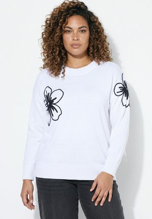 Kvinde med krøllet hår iført en hvid sweater med sorte blomsterdesigns på brystet og ærmet, kombineret med mørke bukser.