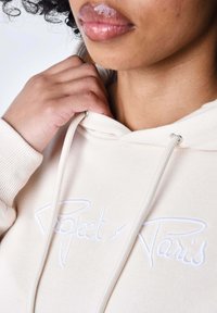 Hoodie beige con una texture morbida, caratterizzato da un logo ricamato bianco "Project x Paris" e occhielli in argento per i lacci.
