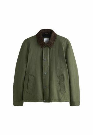 Mann trägt grüne Jacke, marineblau gestreiftes Poloshirt, blaue Jeans und braune Lederschuhe und steht vor beigem Hintergrund.