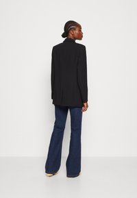 Svart blazer med en strukturert design, enkelt knapp, og hakkede slag, kombinert med flagrende mørkeblå jeans og beige fottøy.