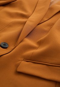 Brun kavaj tillverkad av smidigt tyg med en ensam svart knapp, slag i notch-modell och en framficka på vänster sida.