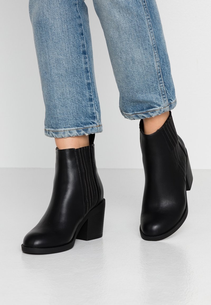Bottines noires en cuir à talons blocs portées avec un jean bleu clair à revers sur un sol et un fond blancs.