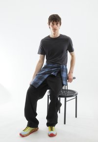 Schwarzes T-Shirt, dunkle Jeans und ein blaues kariertes Hemd, das um die Taille gebunden ist. Bunte Sneakers mit gelben Schnürsenkeln und roten Akzenten. Schwarzer Stuhl.