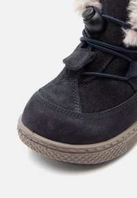 Botte en daim bleu foncé avec une semelle en caoutchouc, des lacets élastiques et une base grise texturée ; présente un doux bord en fourrure autour de la cheville.