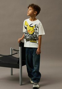 Boy standing next to a black and silver chair wearing a white Pikachu graphic t-shirt, baggy jeans, and sneakers, looking to the side.