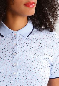 Ljusblå pikétröja med en strukturerad tyg, har en marinblå krage, knappar och ett mönster av små röda, marinblå och svarta hjärtan.