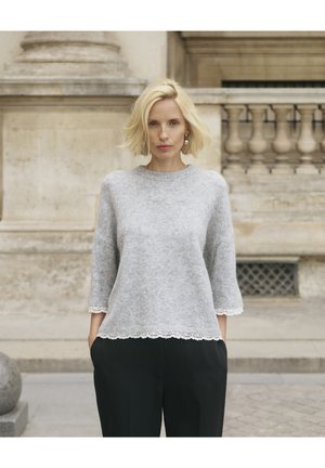 Femeie blondă stă în aer liber purtând un pulover gri deschis cu dantelă la margini și pantaloni negri, pe fundalul unei clădiri de piatră.