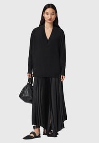 Vrouw in een zwarte oversized trui met diepe V-hals, lange plisseerrok met voorkeurschede, zwarte flats met enkelbandjes, en een zwarte leren handtas.