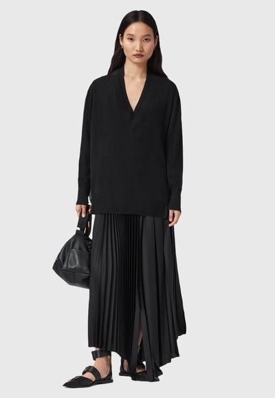 Femme portant un pull oversized noir à décolleté en V profond, une longue jupe plissée avec fente à l'avant, des ballerines noires à bride de cheville, tenant un sac à main en cuir noir.
