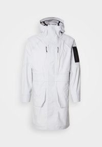 Parka alb impermeabil cu guler înalt, glugă ajustabilă, două buzunare frontale și un buzunar pe mânecă cu fermoar. Are un material elegant, fin.