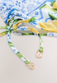 Primo piano di lacci in tessuto con motivo floreale e fibbie metalliche circolari dorate su un indumento stampato blu, verde, giallo e bianco.